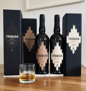 Two bottles of Thomson Whisky with packaging and a glass on a wooden surface.