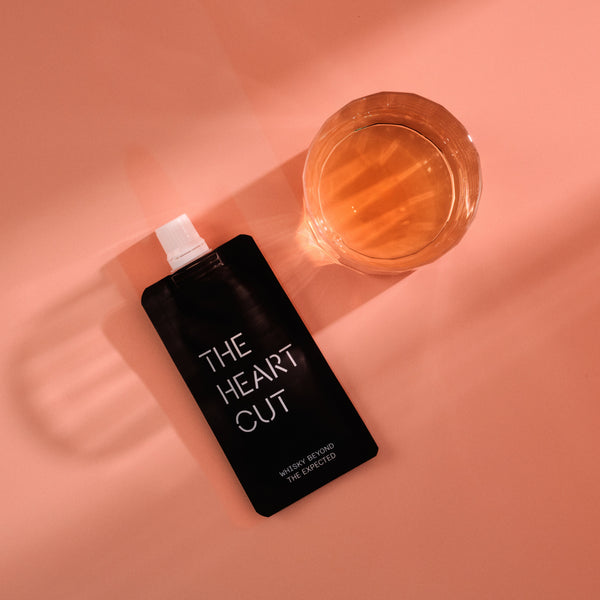 Black bottle labeled 'The Heart Cut' next to a glass of orange liquid on a peach-colored background