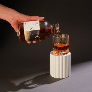 A hand pouring The Heart Cut #23 Boann Distillery single cask Irish whiskey into a crystal rocks glass on a white ribbed pedestal, with dramatic dark background and shadow