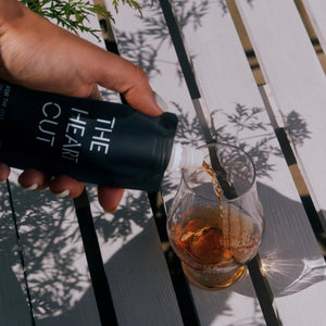 A hand pours a dark liquid from a black bottle labeled 'THE HAIR CUT' into a clear glass containing an amber drink, set on a white slatted wooden surface with leaf shadows.